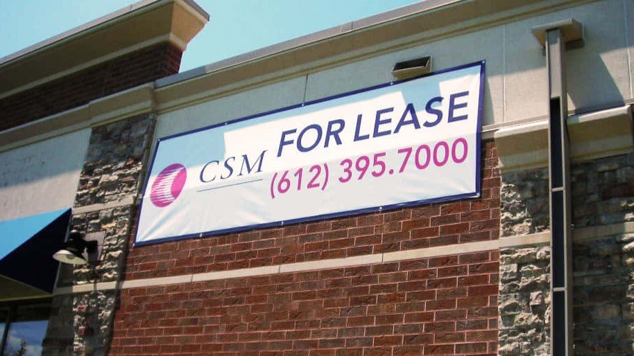 White vinyl "For Lease" banner with contact info mounted on a brick business exterior.